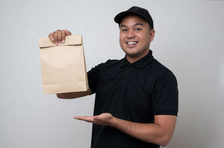 Young smiling asian delivery man in black uniform holding paper bag food delivery on isolated white background.の写真素材