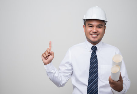 Young handsome asian civil engineer holding paperwork blueprint roll standingpost in studio. Worker...