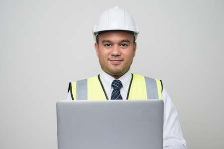 Asian civil engineer operate with laptop to control working in studio. Worker wearing hard hat using laptop computer standing on isolated white background.の写真素材