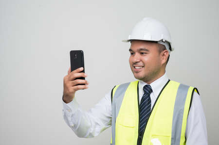 Asian engineer holding mobile phone. Worker wearing hard hat using smartphone standing on isolated...