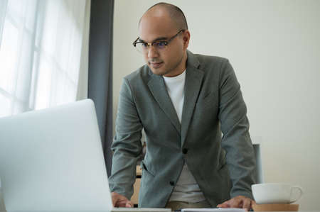 Young asian businessman aged around 30 wearing a grey suit and glasses working in office with...