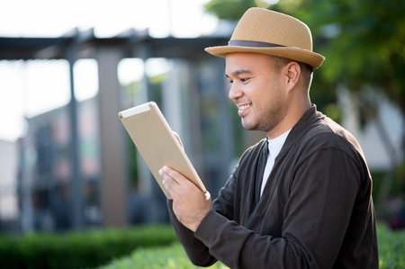 Asian freelancer put on brown hat working outdoor. Office worker or businessman take a tablet for social distancing feel happy at garden area. Outdoor Activity Concept.の写真素材