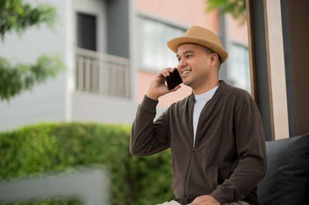 Asian freelancer put on brown hat working outdoor. Office worker or businessman sitting on sofa feel...