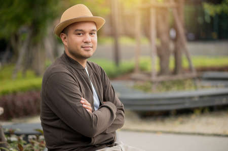 Asian freelancer put on brown hat working outdoor. Office worker or businessman sitting at park area and cross his arm look into camera. Outdoor Activity Concept.の写真素材