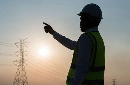 An Asian male engineer works in front of a high-voltage tower pointing the finger. In the sunset the sky is beautifulの写真素材