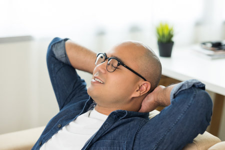 Young asian man Sit and relax in the living room on the sofa. He rested from work raise hands on head and smile.の写真素材