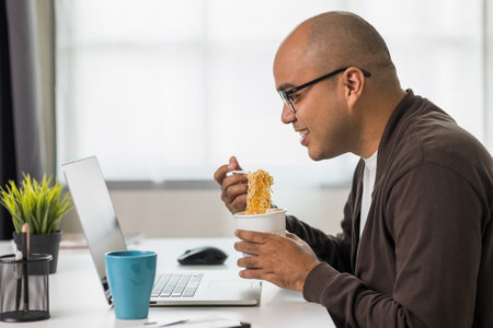 Young asian businessman working with laptop. Indian freelancer eating instant noodle while working. He work at home. He had to hurry and finish his work.の写真素材