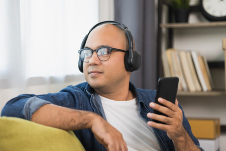 Young asian man wearing headphone and listen the music from smartphone. He in living room sitting on sofa.の写真素材
