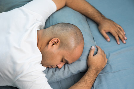 An Asian bald man around the age of 30 in a white T-shirt is sleeping Lying face down on a blue...