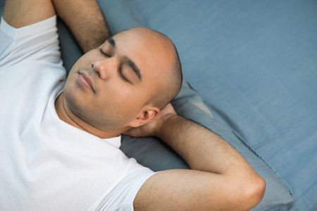An Asian bald man around the age of 30 in a white T-shirt is sleeping on a blue mattress and...