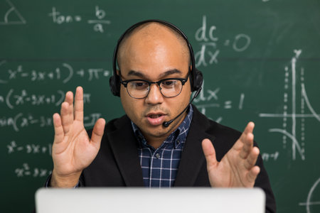 Indian young teacher man sitting wearing headset teaching online video conference live stream by laptop. Asian teacher teaching mathematics class webinar online for students learning.の写真素材