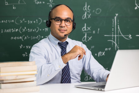 Indian young teacher man sitting wearing headset teaching online video conference live stream by laptop. Asian teacher teaching mathematics class webinar online for students learning.の写真素材