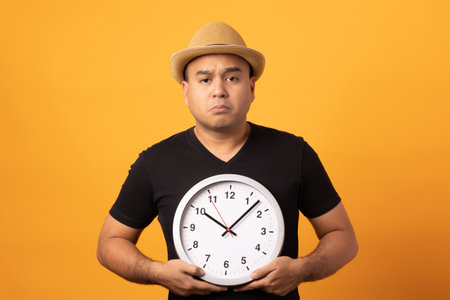 Young asian man wearing hat black shirt holding Big clock standing on isolated yellow background.He...