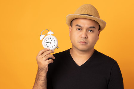 Young asian man wearing hat black shirt holding small alarm clock standing on isolated yellow background. He feeling happy and excitedの写真素材