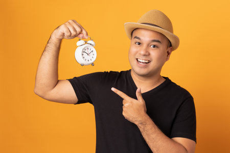Young asian man wearing hat black shirt holding small alarm clock standing on isolated yellow...