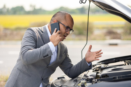 Young asian business has broken down car. He open hood and check the engine. He using smartphone...