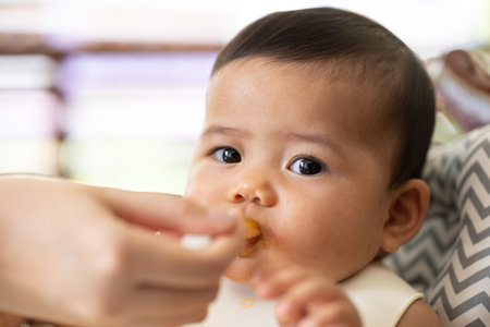 Happy infant baby sits in a child's chair eating first meal.Mom feeds baby.の写真素材