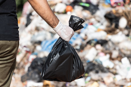 Hand holding a black trash bag in front of a large mountain and a pile of trash. Garbage plastics from industrial and community. World Environment Day.の写真素材