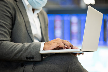 Businessman in airport terminal working with laptop . He wearing mask. He check in and waiting in passenger building.の写真素材