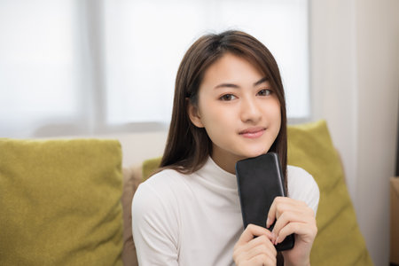 Happy young asian woman looking at screen of smartphone sitting on sofa in living room she shopping online. She very happy. Teenage girl reading news from mobile phone feeling excited.の写真素材