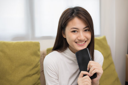 Happy young asian woman looking at screen of smartphone sitting on sofa in living room she shopping online. She very happy. Teenage girl reading news from mobile phone feeling excited.の写真素材