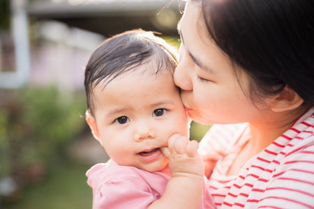Asian mother holding daughter in arms. She held her daughter in her embrace. Parents embracing kissing little baby outdoor with sunny day.の写真素材