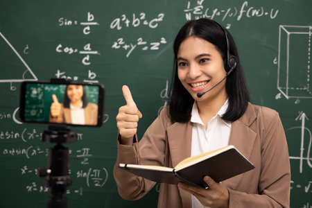 Young asian teacher woman standing looking at camera and video conference with student. Female teacher training the mathematics in classroom from live stream with smartphone online course.の写真素材