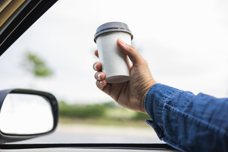 Young man drinking a cup of hot coffee while driving car to travel. Hands holding steering wheel.の写真素材