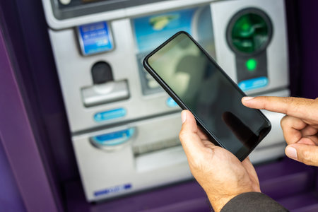 Young businessman standing front of atm machine using smartphone withdraw the money without atm credit card. He ut the pin code into smartphone and take the money.の写真素材