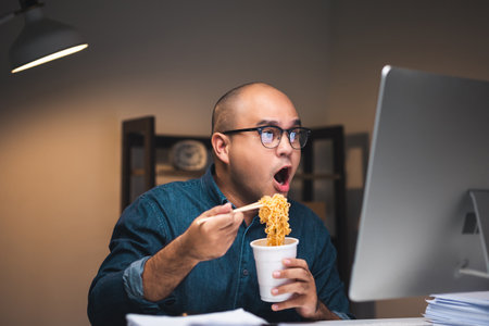 Young asian business freelancer working at late night. He very hungry and eating hot instant noodle and work with computer in the dark office at night.の写真素材