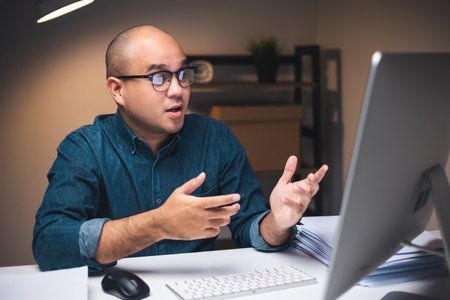 Young asian businessman working with computer video conference in the dark office at night. Attractive Indian man work hard overtime in home with floor lamp ambient warm light late at nightの写真素材