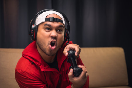 Playing video game. Young asian handsome man sitting on sofa holding joystick in living room. Happiness Streamer Indian man wearing headset playing game online in the darkroom.の写真素材