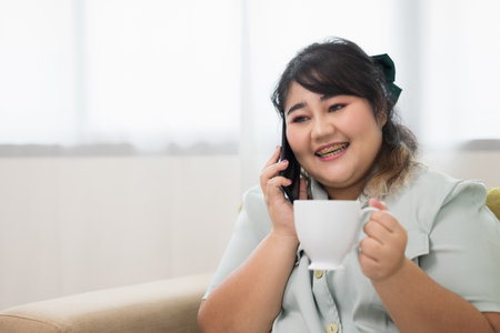 Happy young women's plus sitting on sofa drinking coffee in living room at home. Body positive plus size female lives happily and is proud of herself.の写真素材