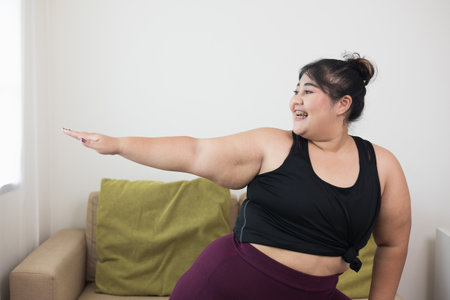 Attractive asian young fitness woman plus size stretching arms warming up before doing yoga at home. Beautiful smiling Female wearing sportswear exercise training yoga in living room.の写真素材
