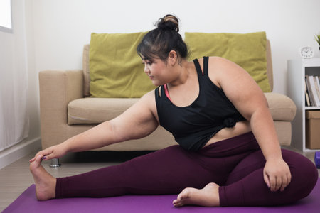 Attractive asian young fitness woman plus size stretching legs warming up before workout at home. Beautiful smiling Female wearing sportswear exercise training yoga in living room.の写真素材