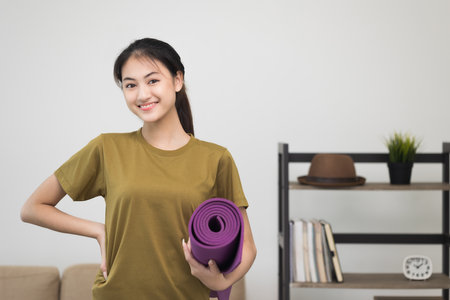 Young beautiful asian woman standing pose smiling to camera holding yoga mat. Cheerfully sporty female workout and exercise wearing sport wear. Charming woman relax after fitness training.の写真素材