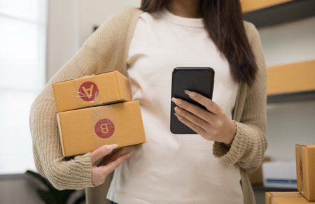 Entrepreneur standing pose holding smartphone and parcel paper cardboard. Female small Business owner with box working at home. Startup freelance people with many parcel on background.の写真素材
