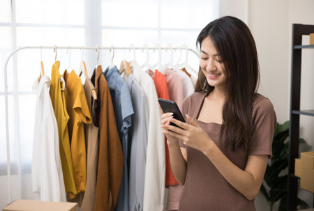 Young beautiful asian woman business owner at fashion store using smartphone check stock on rails clothing standing and smile. Asian female entrepreneur working in clothes shop.の写真素材