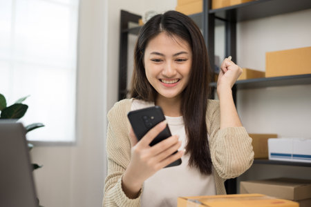 Happy Young asian entrepreneur woman checking product on smartphone. Business woman working SME online at home. Check address on parcel. Small business owner start up freelance concept.の写真素材