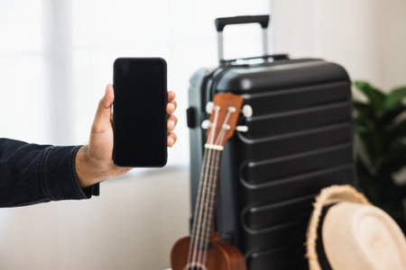 Close up male hands showing blank screen smartphone near trolley bag in the house. Book a trip on smartphone and pay for it. Search for a route to travel by cell phoneの写真素材