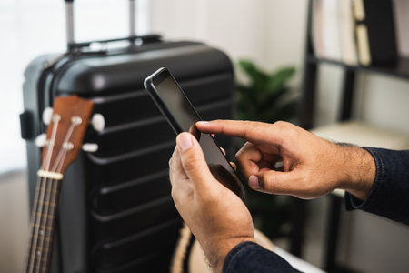 Close up male hands using smartphone near trolley bag in the house. Book a trip on smartphone and pay for it. Search for a route to travel by cell phoneの写真素材
