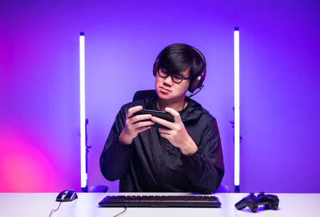 Playing video game on smartphone. Young asian handsome man sitting on chair holding cellphone in living room. Happiness Streamer. Young man wearing headset playing game online in the darkroom.の写真素材