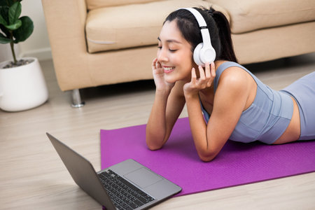 Young beautiful asian woman put on headphone listening music by laptop relax after workout. Lay down on yoga mat. Cheerfully sporty female workout and exercise wearing sport wear at home.の写真素材