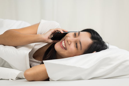 Pretty asian woman using smart phone on bed to social media at home in the morning. Lifestyle of young beautiful female using mobile phone to network internet online on the bedの写真素材