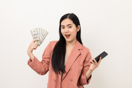 Cheerful young Asian business woman showing lot of money and smartphone. Attractive female with dollar banknotes. Happy asian woman  counting money us dollar on isolated white backgroundの写真素材
