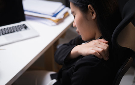 Young businesswoman works late at night in a dimly lit room. she tired and stressed needs rest. She has shoulder neck and back pain. Working hard and overtime too much going to symptom office syndrome.の写真素材