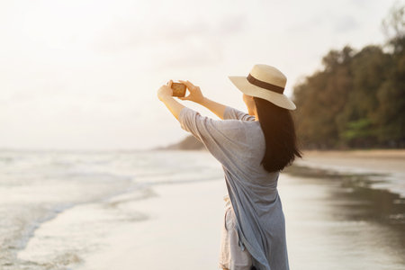 Young asian woman using smartphone to take a photo of seaview while sunset time. Traveler female Relaxing on holiday weekend vacation time. Pick up cell phone to capture the impressionの写真素材