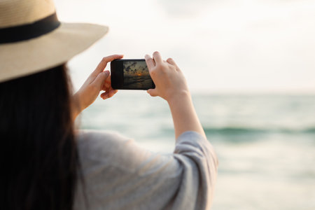 Young asian woman using smartphone to take a photo of seaview while sunset time. Traveler female Relaxing on holiday weekend vacation time. Pick up cell phone to capture the impressionの写真素材