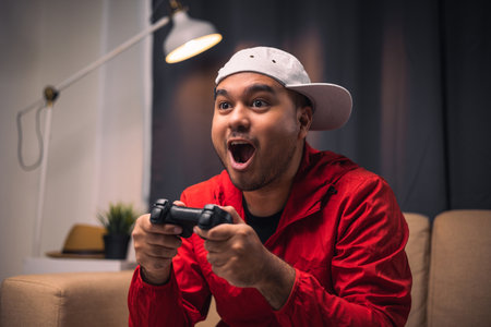 Excited and shocked face of Asian gamer holding joystick playing video game online sitting on sofa...