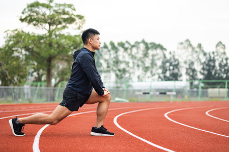 Young asian man wearing sportswear warming up front of sport stadium. Chinese man stretching legs before running on track exercise in the morning. Healthy and athlete lifestyle concept.の写真素材